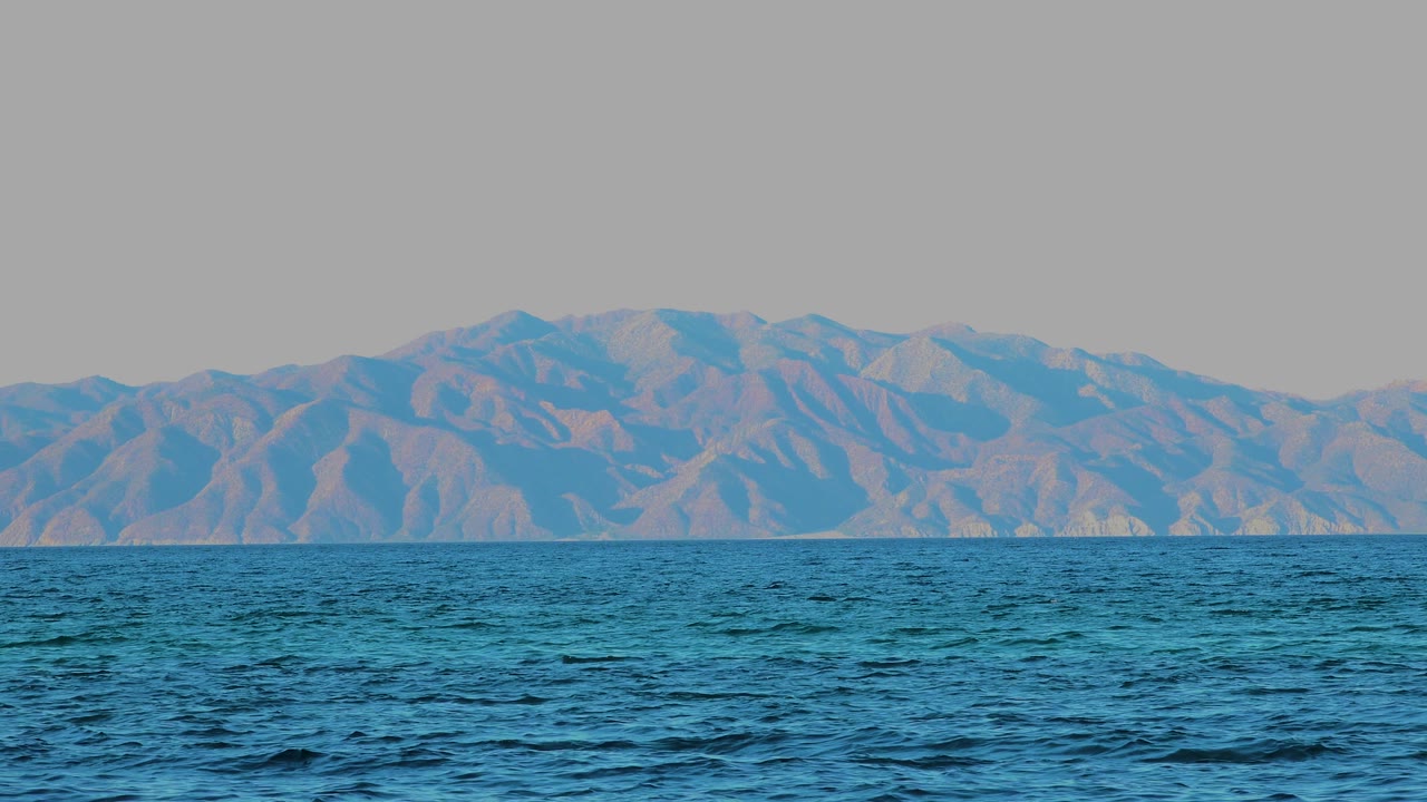 At 0:01, small wave patterns shift across the blue water while the mountain range stays stretched along the horizon in El Sargento La Ventana, Mexico, with no visible people.