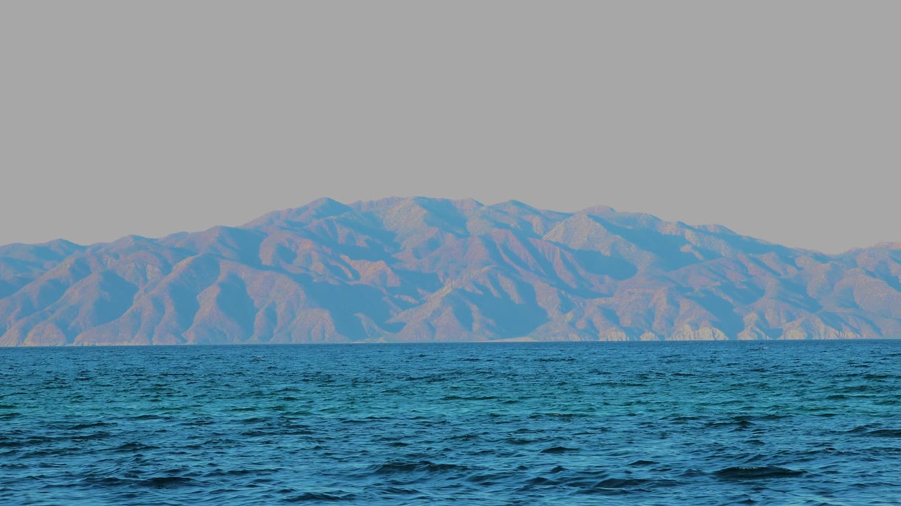 At 0:02, the foreground ocean appears darker and more textured, with tan and blue mountain slopes visible beyond the horizon in El Sargento La Ventana, Mexico and no visible people.