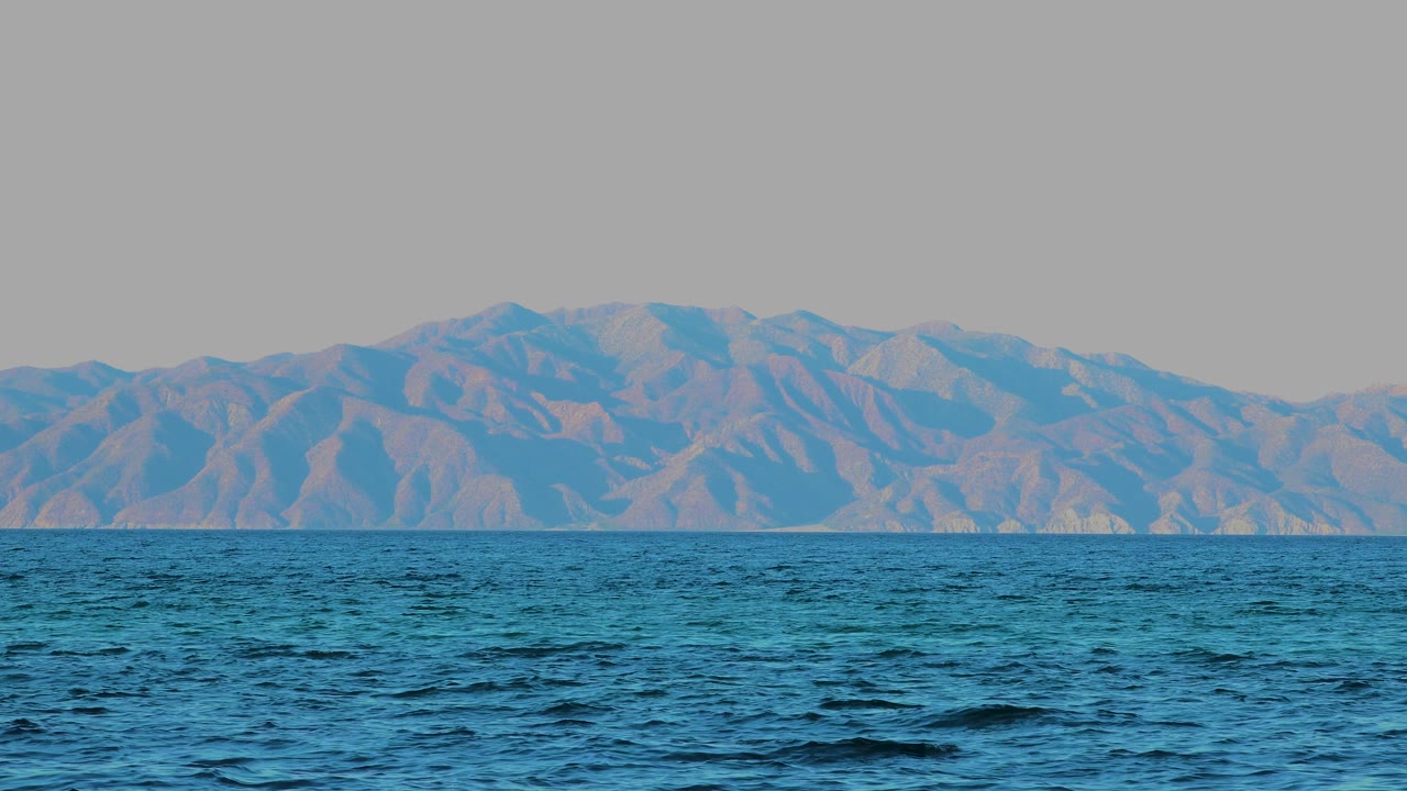 At 0:03, the sea surface carries small dark ripples across the foreground, facing a continuous rugged mountain backdrop in El Sargento La Ventana, Mexico, with no visible people.
