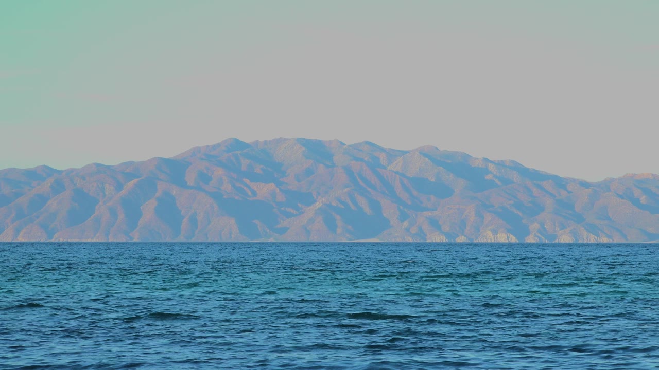 Opening frame of blue water facing distant rolling mountains in El Sargento La Ventana, Mexico, with a pale clear sky and no visible people.