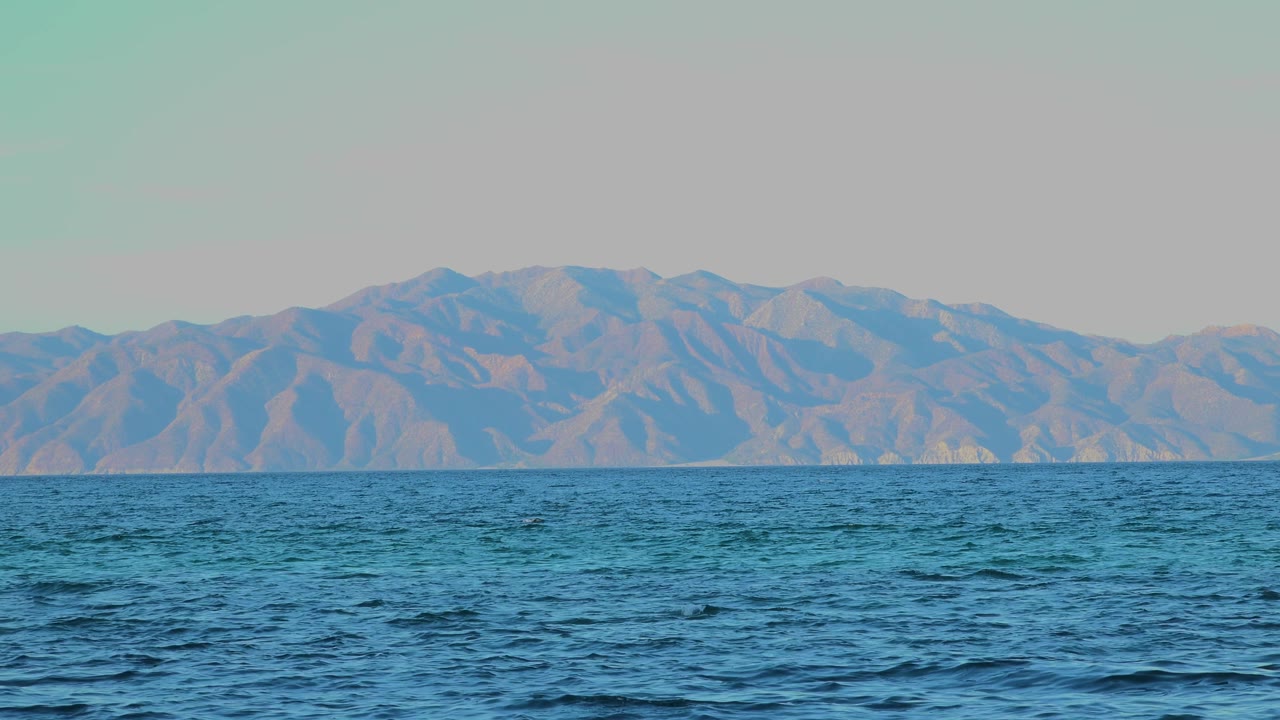 Open blue water runs across the lower frame with pale rolling mountains in the distance and a flat clear sky above in El Sargento La Ventana, Mexico, with no visible people.