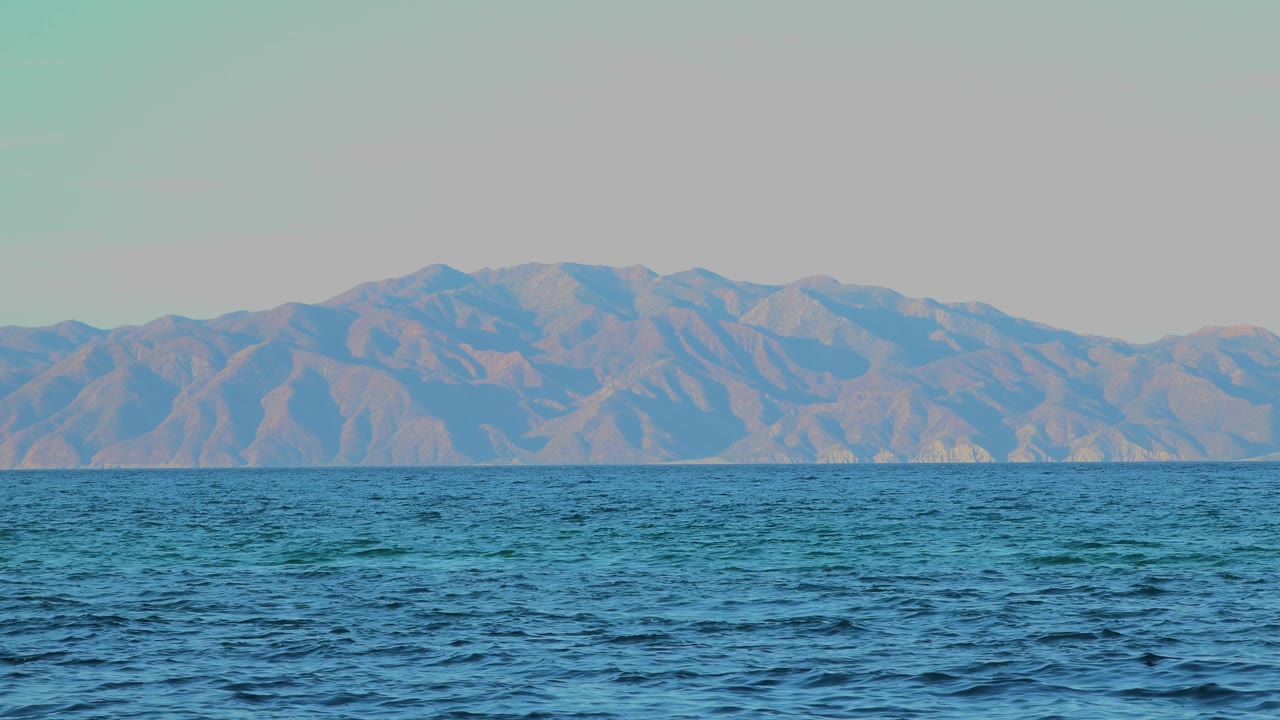Textured blue sea fills the foreground, with a distant mountain range softened by haze across the horizon in El Sargento La Ventana, Mexico, and no visible people.
