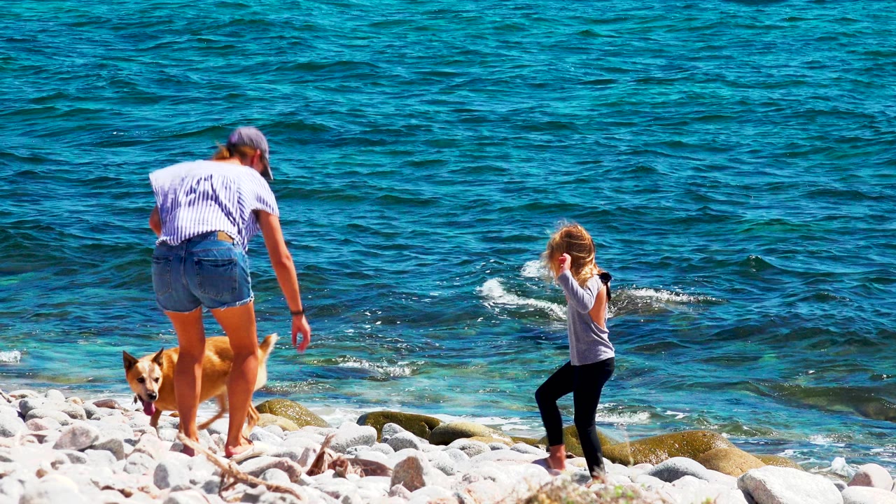 At El Sargento La Ventana, Mexico, a woman bends toward a tan dog while a young girl stands nearby on pale shoreline rocks beside blue water.
