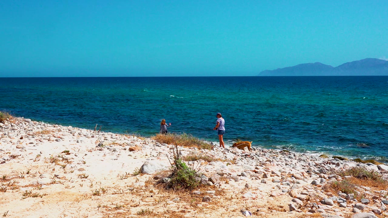 At 0:02 in El Sargento La Ventana, Mexico, two people and a tan dog are positioned farther right on the beach, with small waves and hazy land beyond the blue water.