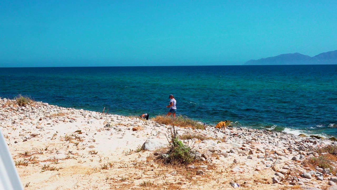 Two people are visible on a pale rocky beach in El Sargento La Ventana, Mexico, with blue green water and distant land along the horizon.