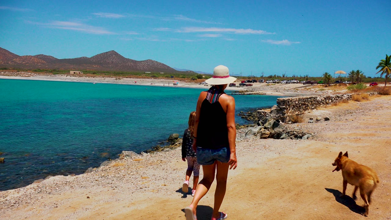 At 0:00 in El Sargento La Ventana, Mexico, a woman in a white sun hat, a child, and a tan dog walk away along a sandy path beside turquoise water.