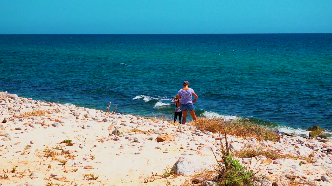 At El Sargento La Ventana, Mexico, a man and child stand near the edge of a pale rocky beach while small waves break below and the horizon runs across the frame.