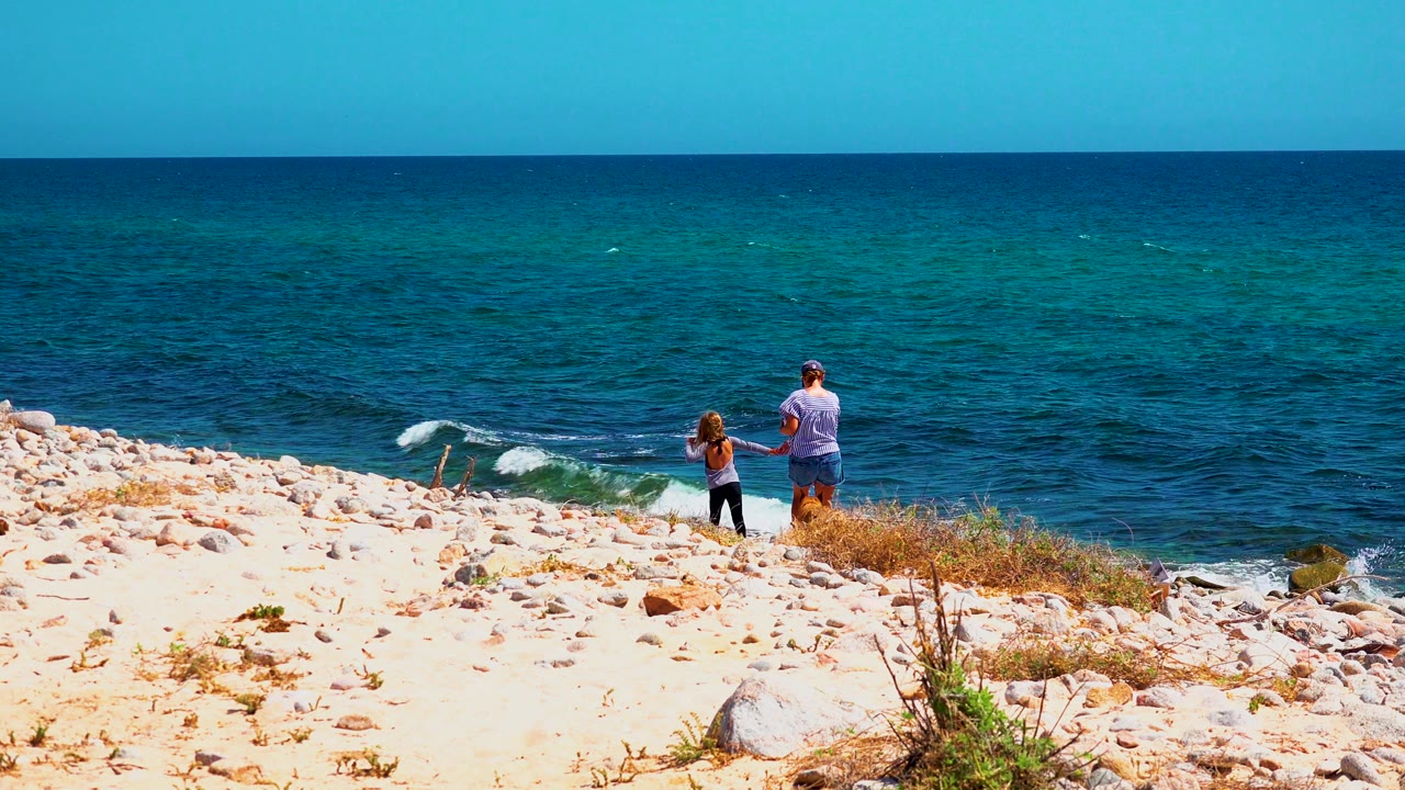 At El Sargento La Ventana, Mexico, the child stands left of the adult on pale sand and stones, with textured blue water filling most of the background.