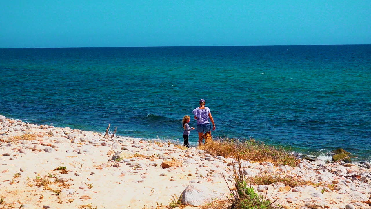 Two people and a small brown dog stand near the rocky shoreline in El Sargento La Ventana, Mexico, with blue-green ocean behind them.