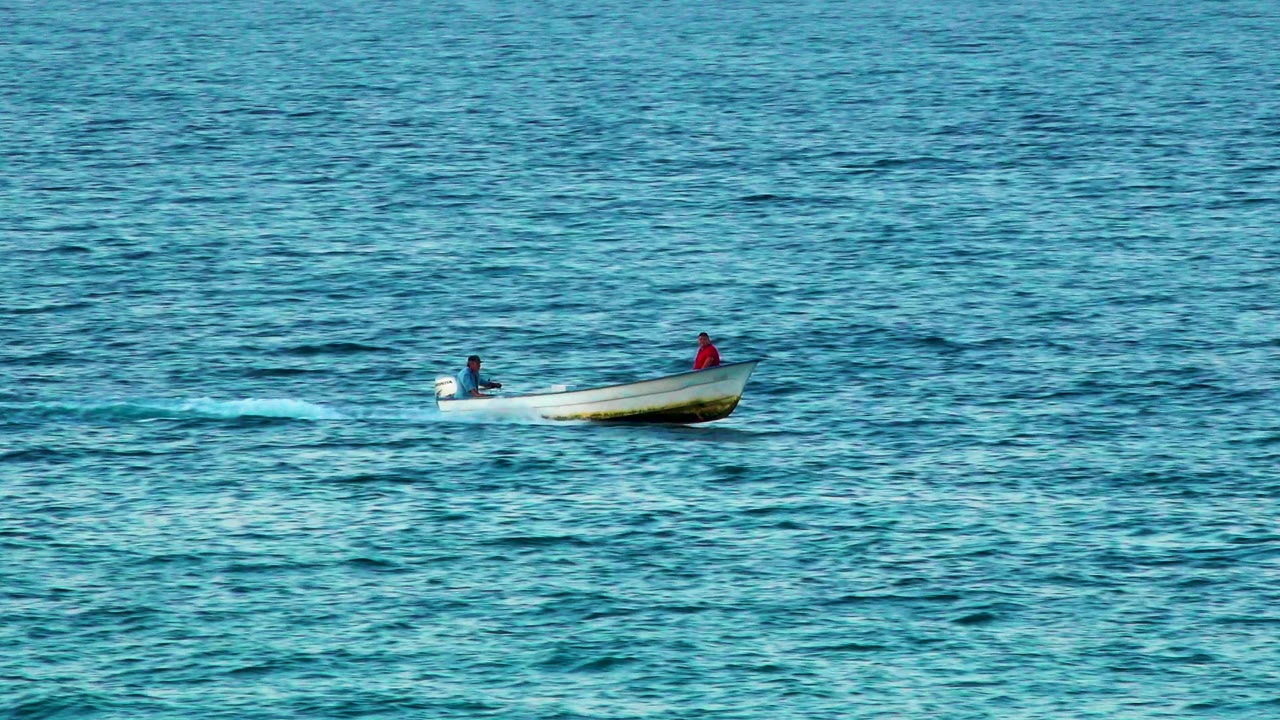 At 0:02, two distant people remain visible in the white boat as it crosses blue-green water with darker ripple lines in El Sargento La Ventana, Mexico.