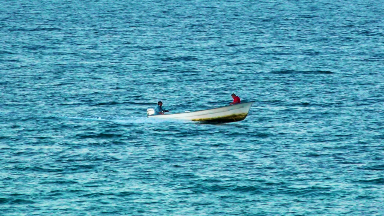 At 0:00 in El Sargento La Ventana, Mexico, a small white motorboat sits left of center on rippled blue water with one seated person near the motor and another in red near the bow.