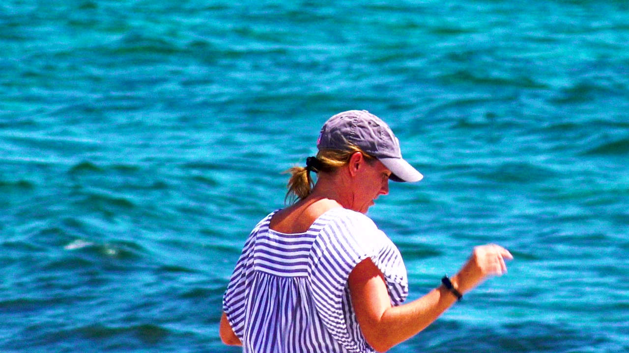 At 0:00, a woman in a gray cap and striped top walks beside bright blue water in El Sargento La Ventana, Mexico, seen from a side angle.