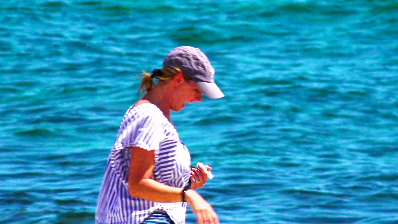 At 0:02, the woman moves farther right in frame, looking down while holding a small item near the blue water in El Sargento La Ventana, Mexico.