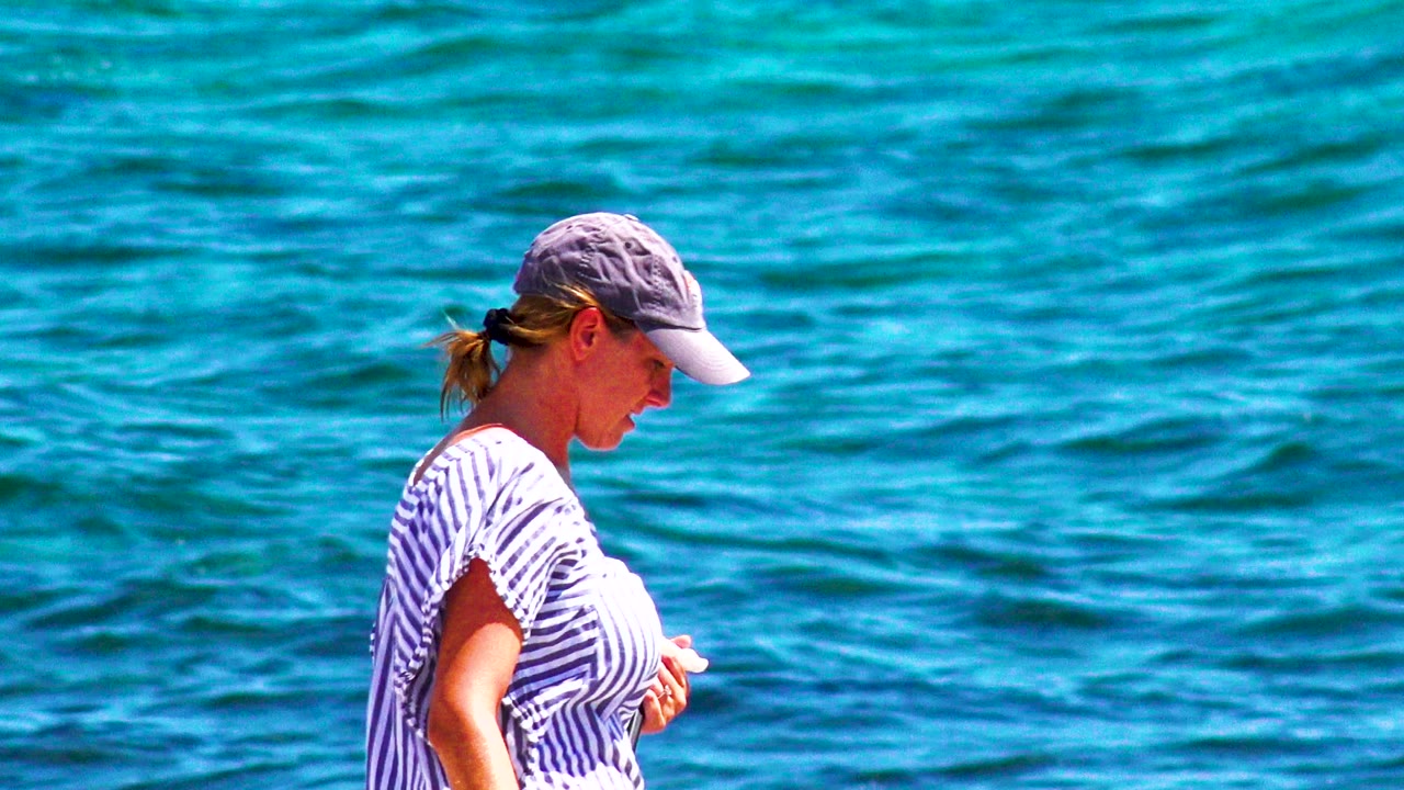 A woman in a gray cap and striped shirt stands in profile near blue ocean water in El Sargento La Ventana, Mexico, with no child visible in this first moment.