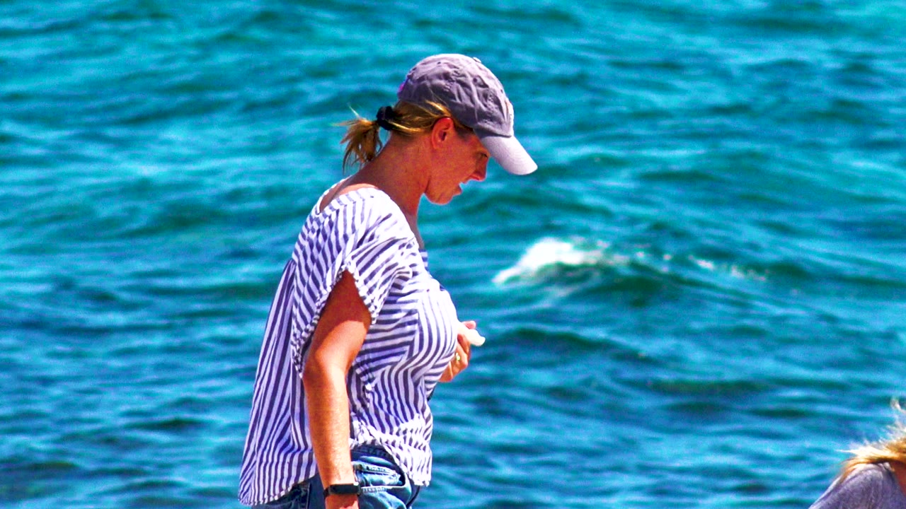 The woman shifts farther right in profile beside bright blue ocean water in El Sargento La Ventana, Mexico, with a partial blond head at the lower edge.