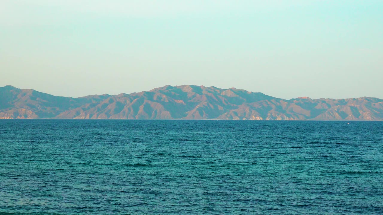 A calm shoreline view holds on blue water and muted brown-blue mountains beneath a soft pale sky, with no visible people.