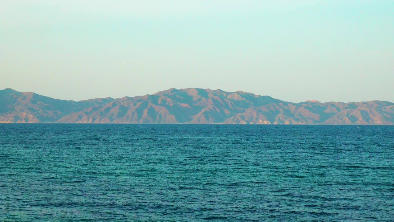 Opening frame of the La Ventana Mexico clip showing blue-green ocean in front of low brown mountains, with pale sky above and no visible people.