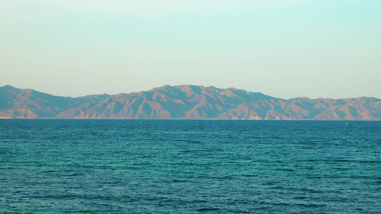 At 0:01, textured ocean water fills the foreground while the distant La Ventana area mountains remain centered beneath a clear pale sky, with no visible people.