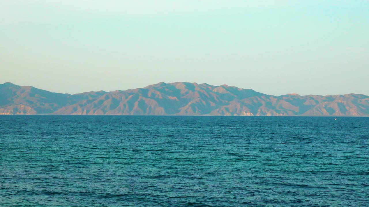 At 0:02, the wide coastal view shows open blue water across the lower frame and a long brown mountain line in the distance, with no visible people or boats.