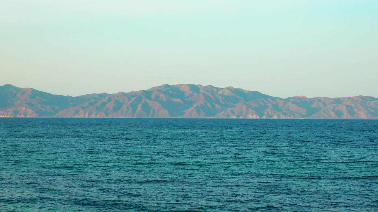 At 0:03, rippling blue-green sea occupies most of the frame with softly lit mountains stretching across the background and no visible people.