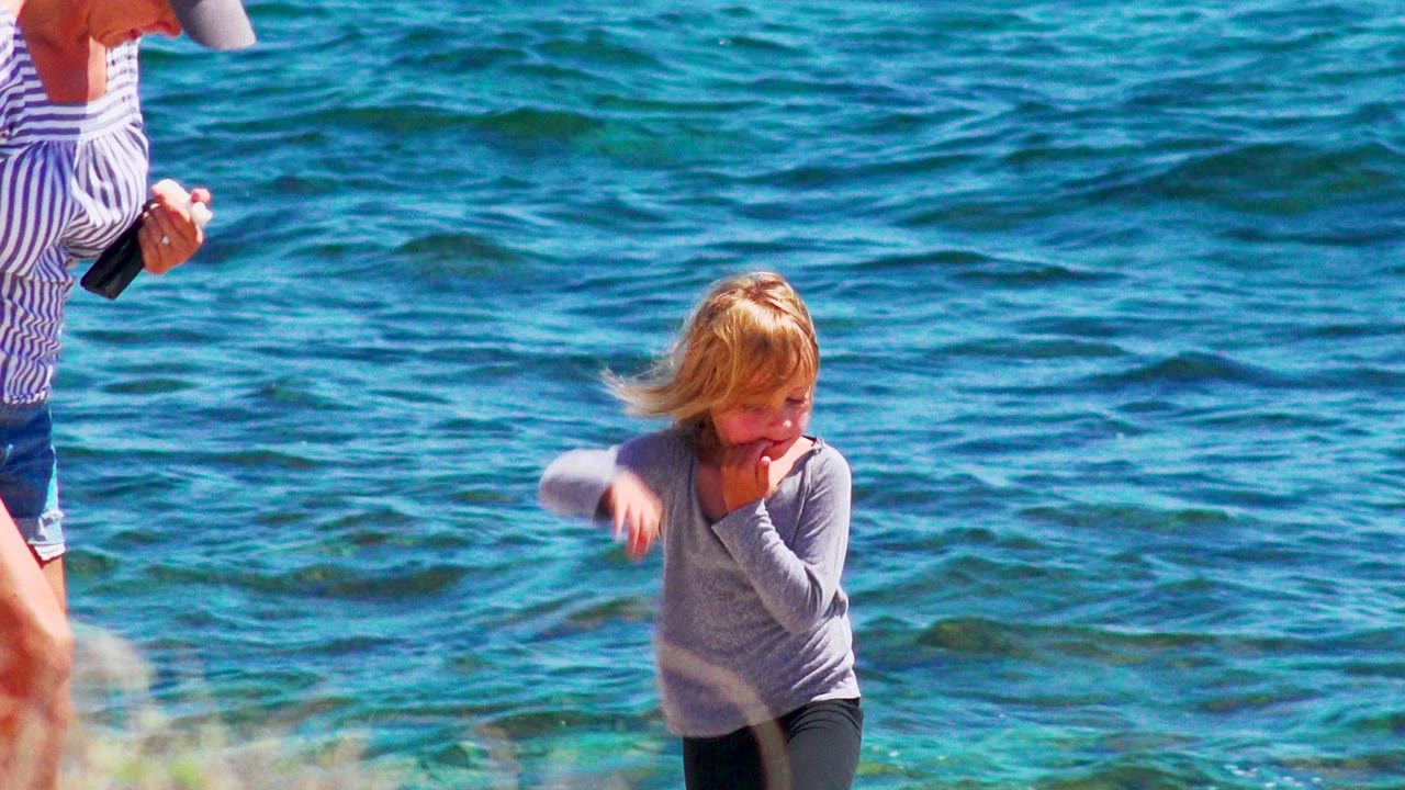 At 0:00 in El Sargento La Ventana, Mexico, a blond child in a gray top walks near bright blue water while a cropped adult stands at the left edge.