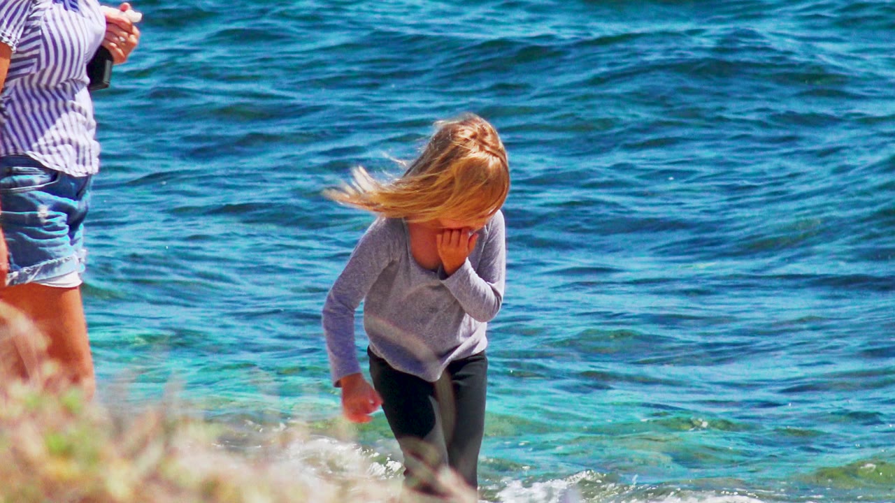 At 0:02 in El Sargento La Ventana, Mexico, the child bends near the waterline with windblown hair, small waves, and the cropped adult's legs at the left edge.