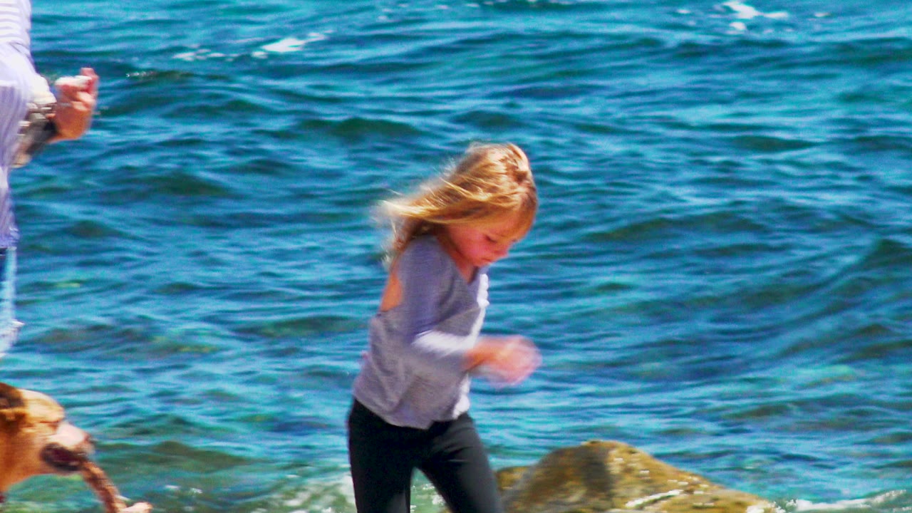 Blonde child in a long sleeve top walks along the El Sargento La Ventana shoreline as blue water fills the background, with a partial adult arm and dog at the frame edges.