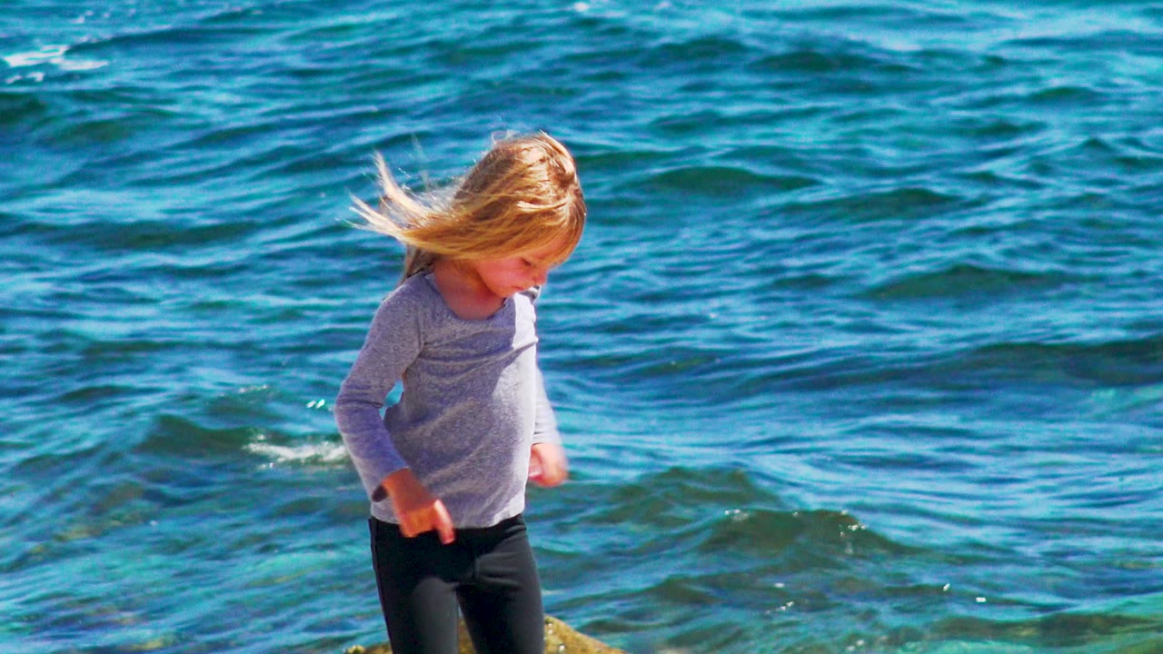 Blonde child in a long sleeve top steps along the rocky El Sargento La Ventana shore while blue-green waves ripple behind her and wind pushes her hair forward.