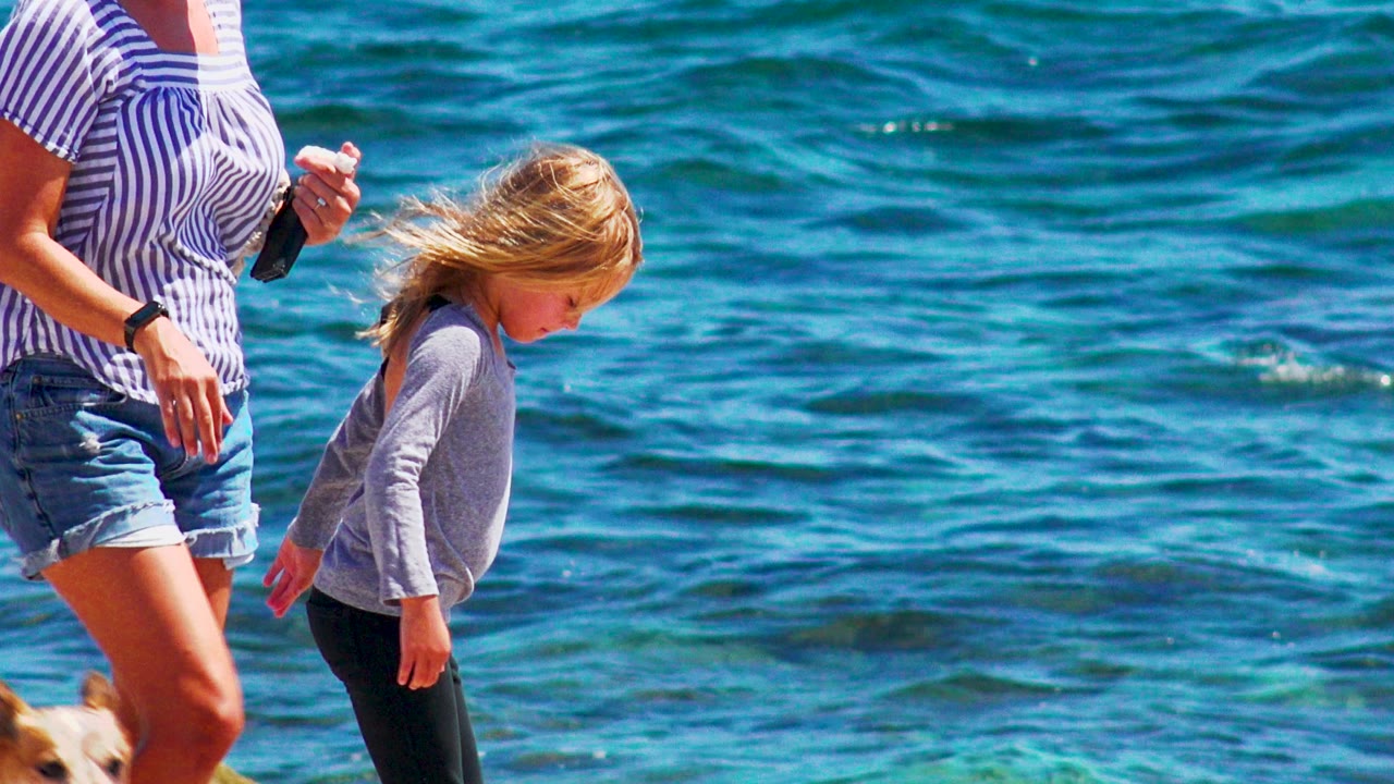 Woman holding a phone walks near a child in front of turquoise water.