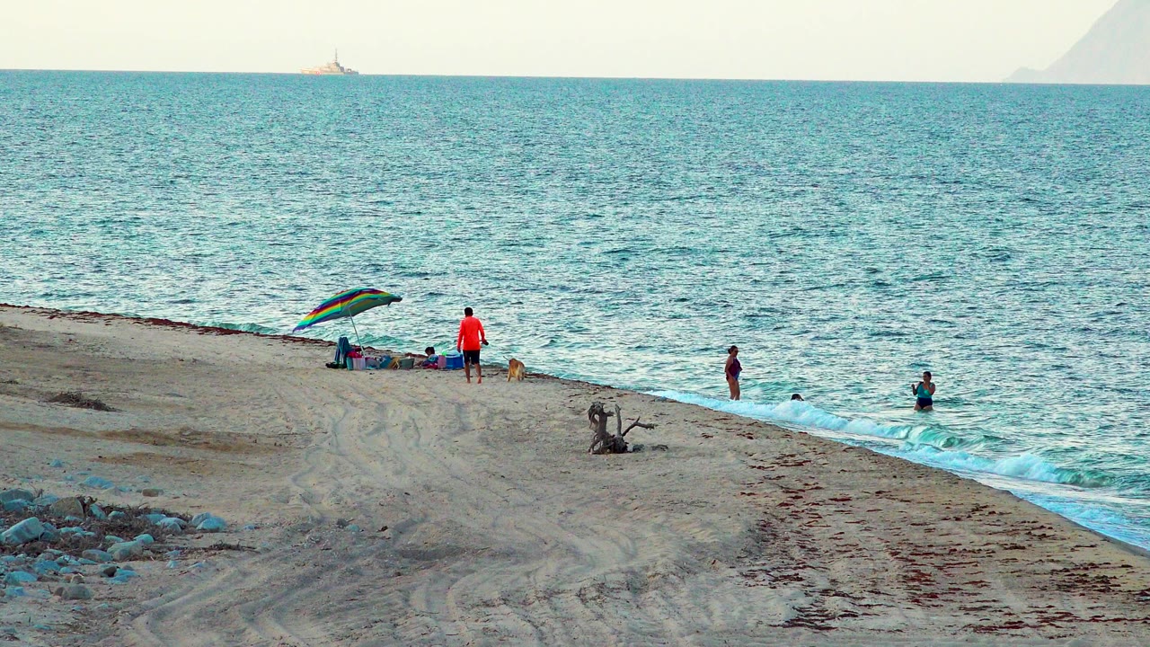 Wide view of El Sargento La Ventana, Mexico beach with people near the surf, a dog by a person in a red top, a rainbow umbrella, and blue-green water beyond.