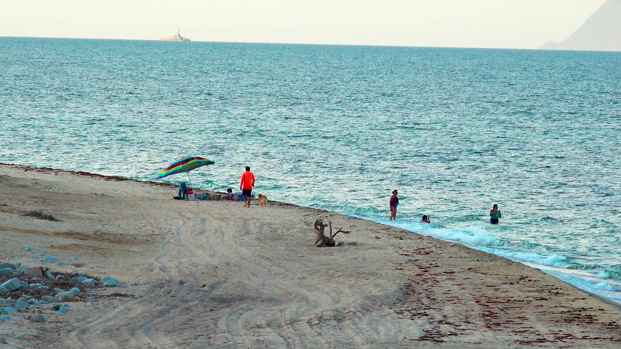 Wide El Sargento La Ventana shoreline with waders in the surf, a person and dog beside the rainbow umbrella, and pale haze over the distant water.