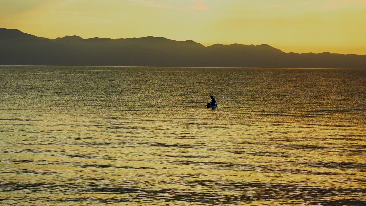 A distant silhouetted rower sits left of center on rippled water in El Sargento La Ventana, Mexico, with dark mountains and yellow evening light behind them.