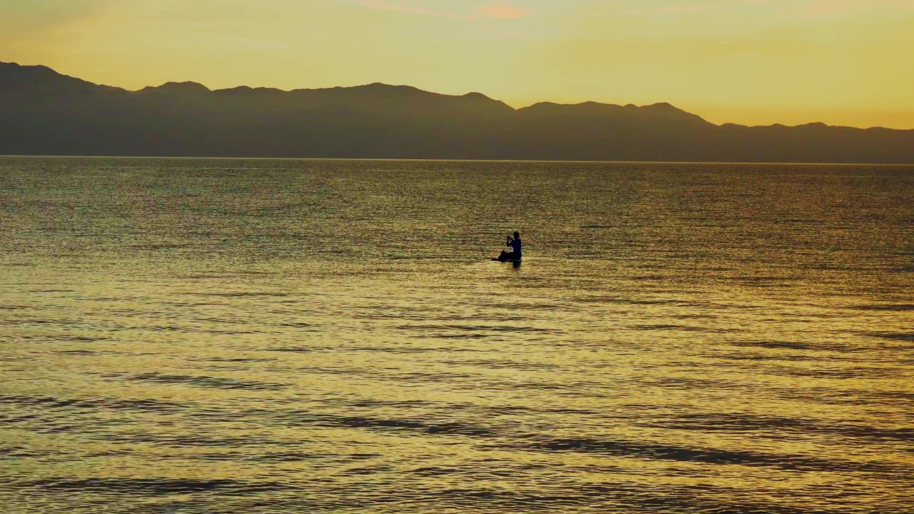 A small silhouetted rower sits left of center on golden ocean water in La Ventana, Mexico, with dark mountains along the horizon.