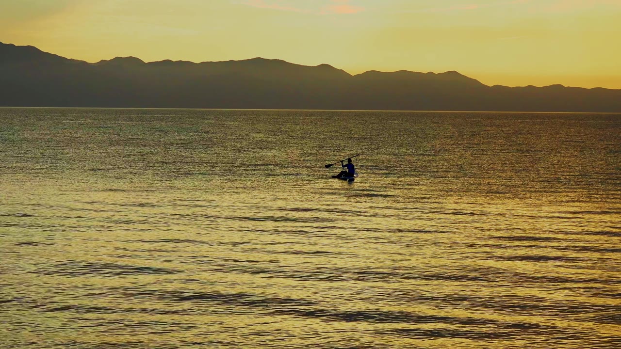 A distant rower and small boat sit right of center on the ocean, with warm light on the water and a dark mountain ridge in the background.