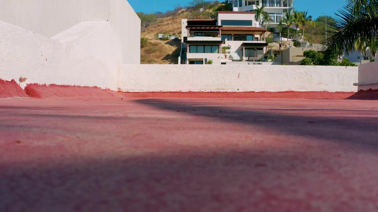 Los Cabos frame with a low red paved foreground, a white stucco wall, hillside modern homes, palm fronds, dry brush, blue sky, and no visible people.