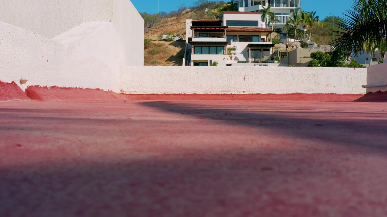 A low exterior composition in Los Cabos shows red paving, a white stucco wall, hillside houses, dry vegetation, palm leaves, and no visible people.