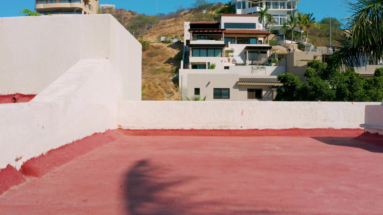 Opening view of a red rooftop surface and white parapet in Los Cabos, with hillside houses, trees, and blue sky beyond, no visible people.