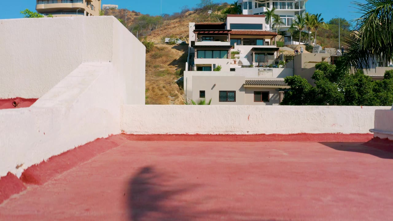 A white rooftop wall dominates the lower frame in Los Cabos while modern homes, palm leaves, dry hillside, and clear sky fill the background, no visible people.