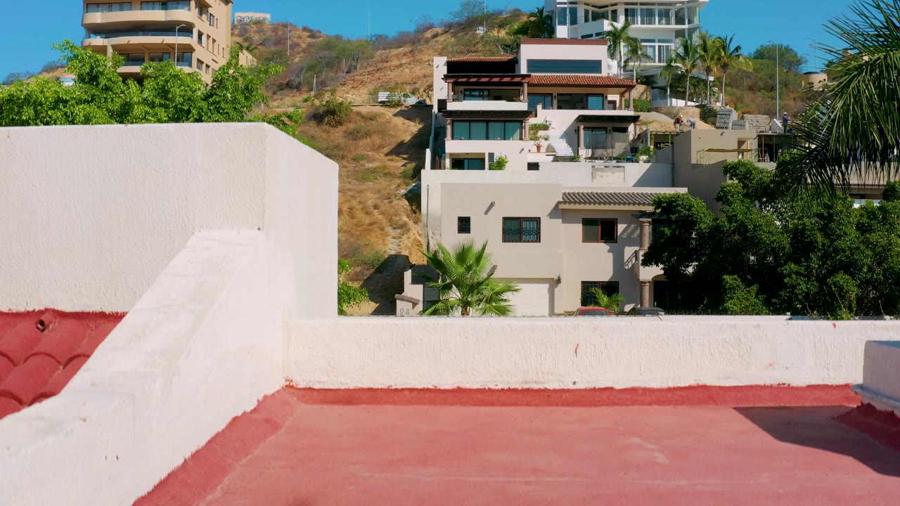 Los Cabos hillside residences appear above a white parapet and red roof edge, with dense green trees and palm foliage on the right, no visible people.