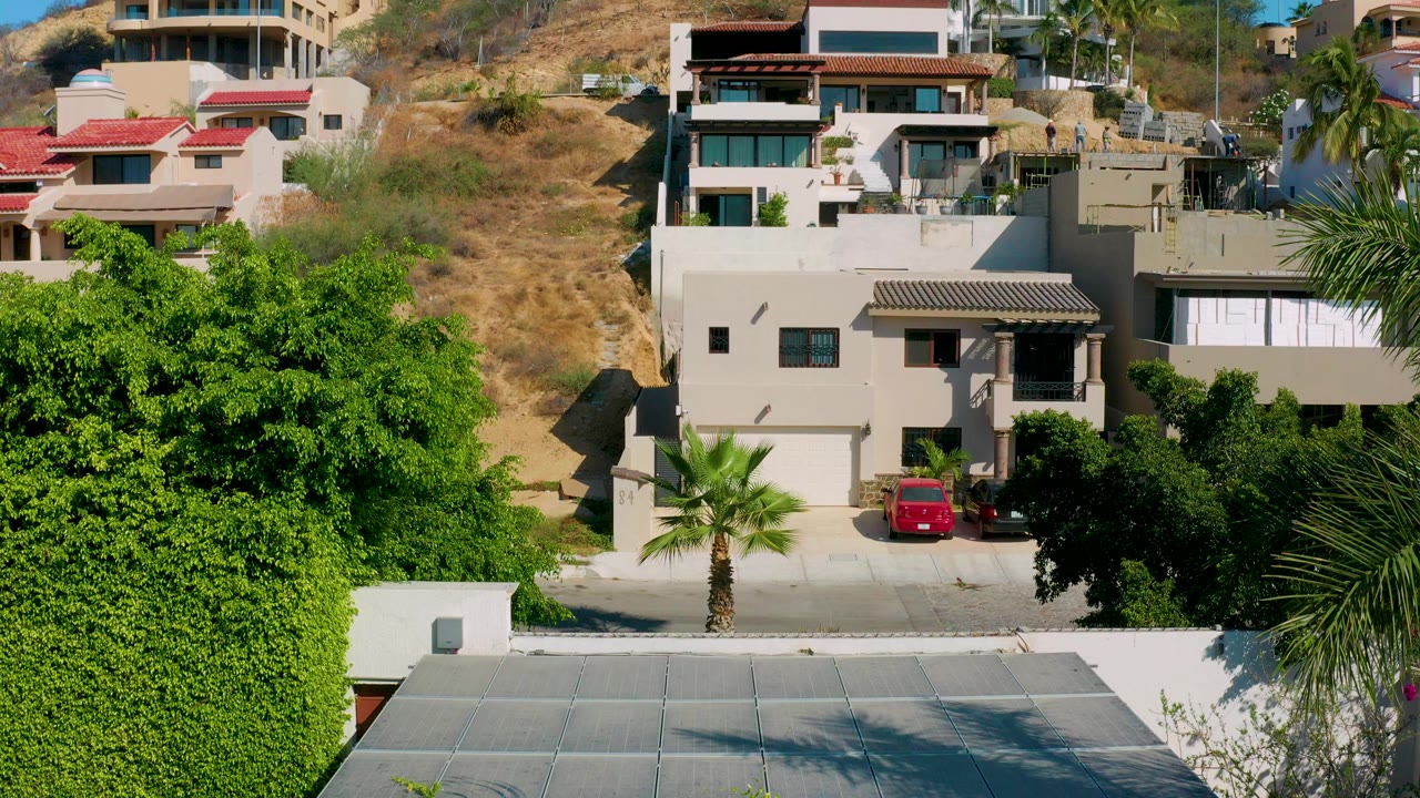 Elevated Los Cabos view with a large green tree in the foreground, a road, parked cars, and modern hillside villas in bright sun.