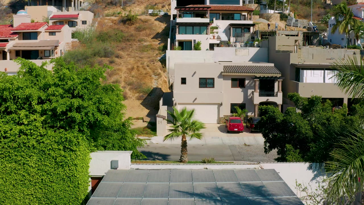 Modern stucco villas in Los Cabos appear above a quiet road, with a red parked car, palms, and dry hillside vegetation behind them.