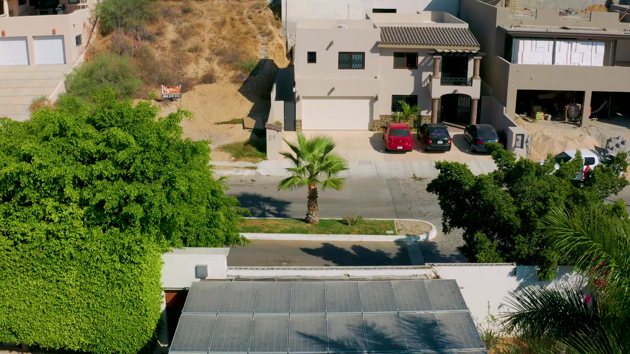 Aerial frame in Los Cabos, Mexico, with a tan house, three parked cars in a driveway, a dry slope behind the homes, and thick green trees in the foreground with no visible people.