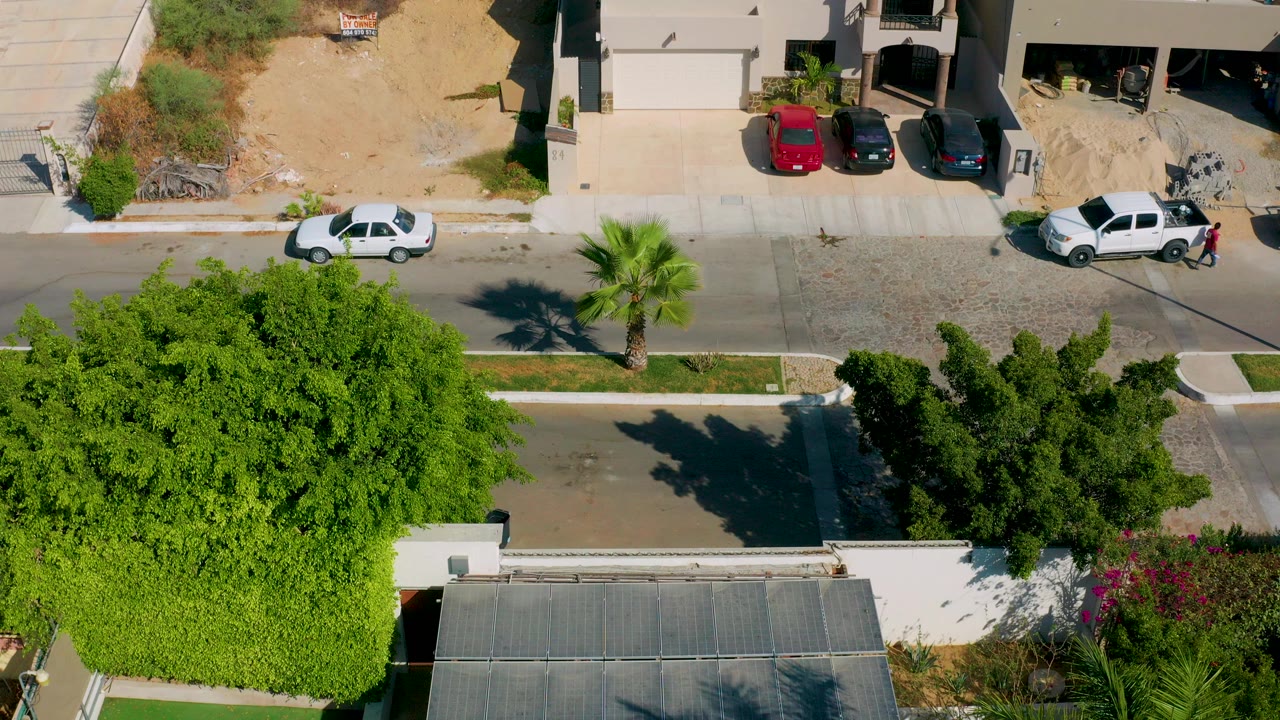 Overhead drone frame in Los Cabos, Mexico showing a quiet residential street, parked white car, house driveways, dense green trees, and no visible people.