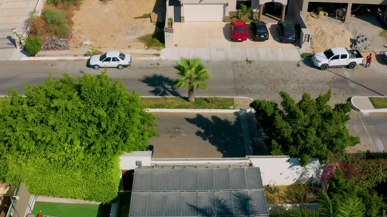 Top-down Los Cabos residential frame showing a white pickup near a driveway, parked cars across the street, green trees, sidewalks, and no visible people.