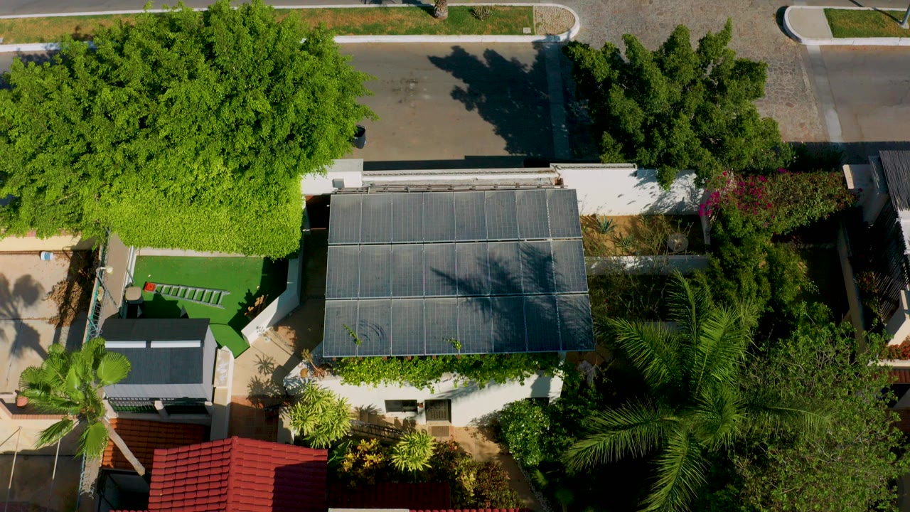 Overhead view in Los Cabos, Mexico showing a modern roof, bright green yard, palms, roadside pavement, and no visible people.