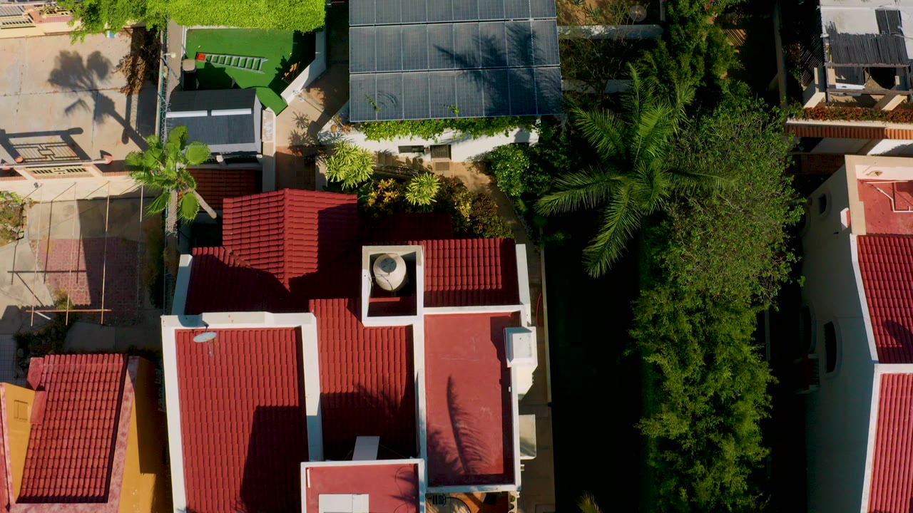 Top-down aerial frame of red-roofed houses, white walls, paved patios, and dense green palms in Los Cabos, Mexico, with no visible people.