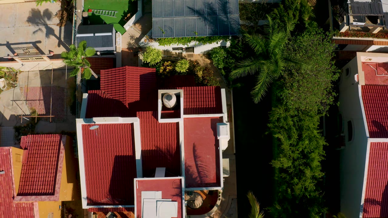 Aerial frame of adjacent homes in Los Cabos, Mexico, with red rooftops, tan patio areas, palm shadows, and a narrow band of dense trees, with no visible people.