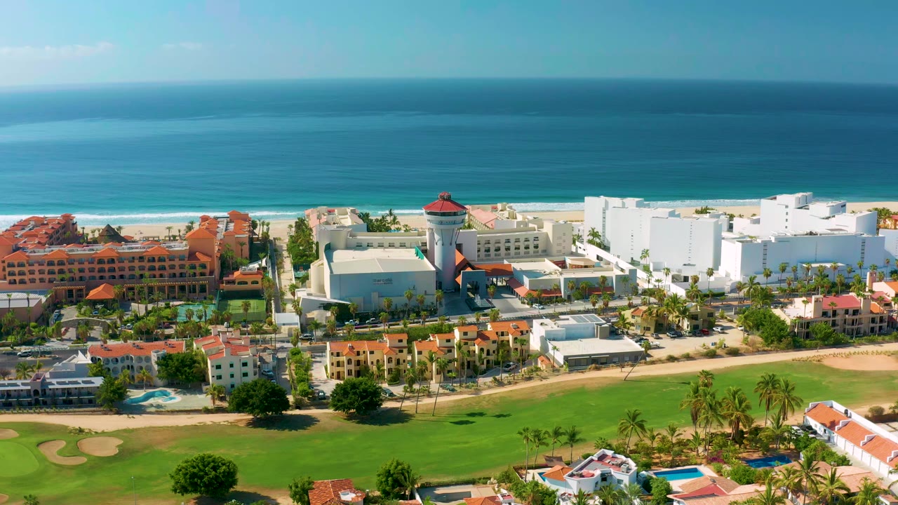 Opening aerial frame over Los Cabos, Mexico, with green golf fairways in front, resort buildings near the beach, blue ocean beyond, and no visible people.