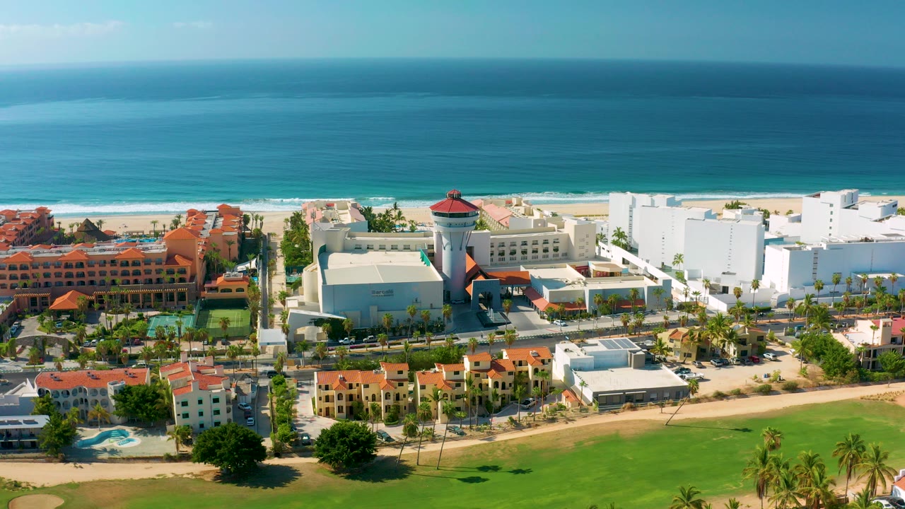 A high aerial frame over Los Cabos shows white and terracotta buildings beside a sandy beach and blue ocean, with no visible people.