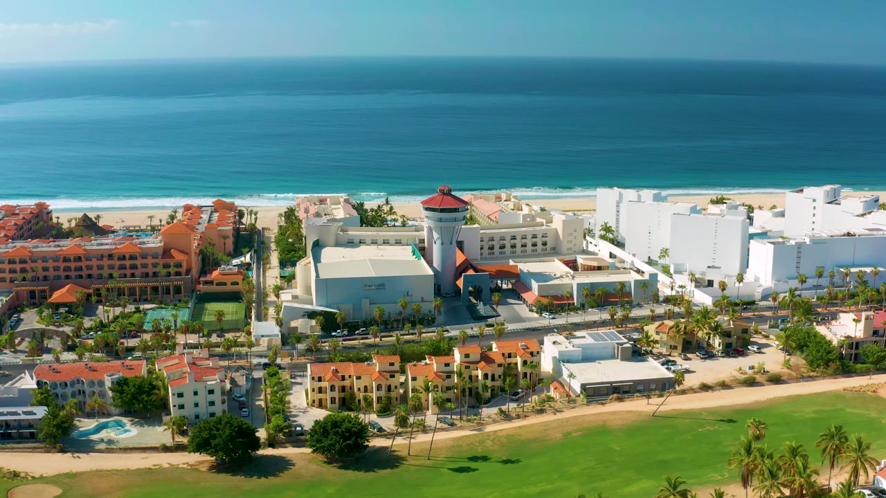 The Los Cabos aerial holds over beachfront buildings, palm-lined roads, pools, and a broad band of turquoise water, with no visible people.